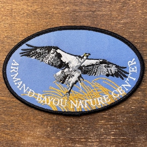 Puede incluir: Parche ovalado con borde negro, que representa un águila pescadora en vuelo sobre juncos dorados sobre un fondo azul claro. El texto "ARMAND BAYOU NATURE CENTER" está curvado alrededor del diseño.
