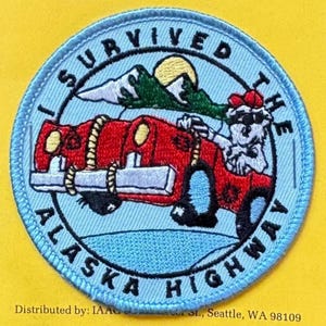 Könnte beinhalten: Gestickter Aufnäher mit hellblauem Rand und dem Text "I SURVIVED THE ALASKA HIGHWAY". Der Aufnäher zeigt ein rotes Auto, das von einem Skelett gefahren wird, Berge und eine Sonne.