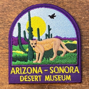 May include: Embroidered patch featuring a cougar in a desert landscape with cacti, a yellow sun, and a bird. The patch has a purple border and the text "ARIZONA - SONORA DESERT MUSEUM" in yellow.