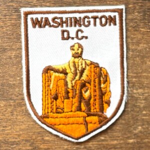 Könnte beinhalten: Gestickter Aufnäher mit dem Text "WASHINGTON D.C." und einem Bild des Lincoln Memorials. Der Aufnäher hat einen weißen Hintergrund mit braunen und orangenen Details und einen braunen Rand.