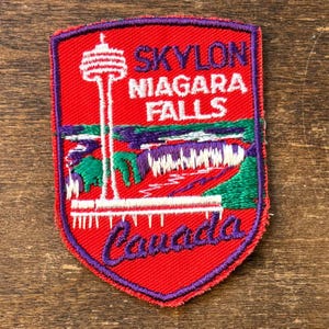 Peut inclure: Écusson brodé représentant la tour Skylon et les chutes du Niagara, avec le texte "Skylon Niagara Falls Canada". L'écusson a un fond rouge avec des accents violets et verts, et une tour blanche.