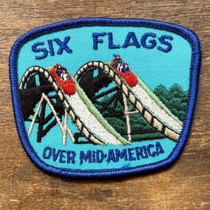 以下が含まれることがあります： 青い縁と背景の刺繍パッチで、ジェットコースターのデザインが特徴です。パッチには青い文字で「SIX FLAGS OVER MID-AMERICA」と書かれています。ジェットコースターの車は赤色で、トラックは黒と白です。