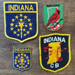 Könnte beinhalten: Vier gestickte Aufnäher mit dem Wort "INDIANA". Einer ist marineblau mit goldenen Sternen, ein anderer ist grün mit einem roten Kardinal, und ein dritter ist schwarz mit einer gelben Karte von Indiana und roten Blitzen. Der vierte ist blau mit goldenen Sternen.