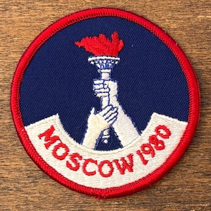 May include: Embroidered patch featuring a torch held by two hands, with a red flame atop. The patch has a blue background, a red border, and the text "MOSCOW 1980" in white.
