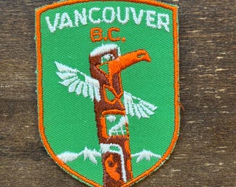 Puede incluir: Un parche bordado verde y naranja con el texto "Vancouver B.C." y un tótem estilizado con un pájaro en la parte superior.