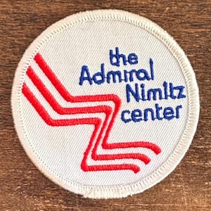May include: A circular white patch with the text "the Admiral Nimitz center" in blue and a red and white zig-zag design. The patch has a white stitched border and is likely an embroidered emblem.