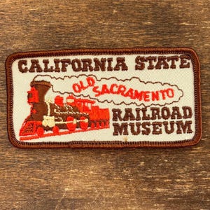 Könnte beinhalten: Ein rechteckiger gestickter Aufnäher mit einer braun-roten Dampflokomotive. Der Text CALIFORNIA STATE OLD SACRAMENTO RAILROAD MUSEUM ist in braunen und roten Buchstaben auf cremefarbenem Grund mit braunem Rand gestickt.
