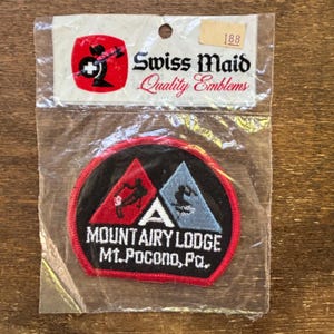 Può includere: Una toppa ricamata in rosso, bianco e blu con il testo "Mountairy Lodge Mt. Pocono, Pa." e due sciatori al centro. La toppa è confezionata in un sacchetto di plastica trasparente con un'etichetta bianca che recita "Swiss Maid Quality Emblems 188".