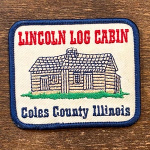 Könnte beinhalten: Gestickter Aufnäher mit einer Blockhütten-Illustration. Der Aufnäher hat einen weißen Hintergrund und einen blauen Rand. Der Text in Rot und Blau lautet "LINCOLN LOG CABIN" und "Coles County Illinois". Die Hütte ist beige mit blauem Dach und grünem Gras.