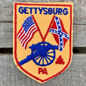 Könnte beinhalten: Ein gelber gestickter Aufnäher mit rotem Rand. Der Aufnäher zeigt den Text "GETTYSBURG" in blauen Buchstaben, zwei Flaggen, eine Kanone und die Buchstaben "PA". Die Flaggen sind die amerikanische Flagge und die Flagge der Konföderierten. Die Kanone ist blau mit einem weißen Rad. Der Aufnäher ist ein Vintage-Souvenir.