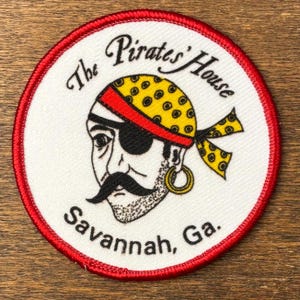 May include: Circular patch with a red border and white center. The patch features a pirate with a black eye patch, a yellow and black polka-dot bandana, and a gold earring. Text reads "The Pirates' House Savannah, Ga."