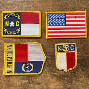 May include: Four embroidered patches featuring flags and symbols. One patch depicts the American flag, while others showcase the North Carolina state flag and symbols. The patches are in various shapes and sizes, with vibrant colors and detailed stitching.