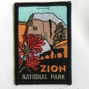 May include: Embroidered patch featuring a landscape scene from Zion National Park. The design includes a mountain, red flowers, and a deer silhouette. The text "ZION NATIONAL PARK" is stitched in orange and green.