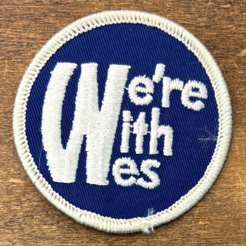 Puede incluir: Un parche bordado circular con un fondo azul profundo y un borde cosido en blanco. El texto "We're with Wes" est&aacute; bordado en blanco, con una "W" may&uacute;scula grande seguida por las letras restantes en una fuente m&aacute;s peque&ntilde;a y apilada.