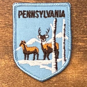 以下が含まれることがあります： 水色の背景にダークブラウンの文字で「PENNSYLVANIA」と刺繍された盾形のワッペン。雪景色の中に立つ茶色の鹿と、それより小さな茶色の動物、そして白い木々がデザインされています。