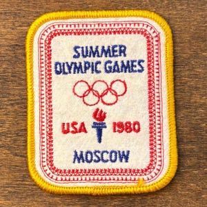 May include: Embroidered patch with a yellow border and a white center. The patch features the text "SUMMER OLYMPIC GAMES" in blue, the Olympic rings, and "USA 1980 MOSCOW" in red and blue. The patch has a red stitched border.