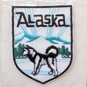 Pode incluir: Etiqueta branca bordada com um cão husky preto e branco, uma cordilheira azul e a palavra "Alaska" em letras pretas.