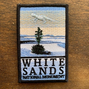 May include: Embroidered patch featuring a landscape scene with a lizard in the sky, a tree, and the text "WHITE SANDS NATIONAL MONUMENT." The patch has a black border and is rectangular in shape.