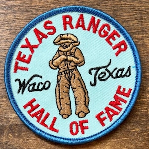 May include: Round patch with a light blue background and a blue border. The patch features a brown illustration of a person in a cowboy outfit. The words "TEXAS RANGER" and "HALL OF FAME" are in red, and "Waco Texas" is in black.