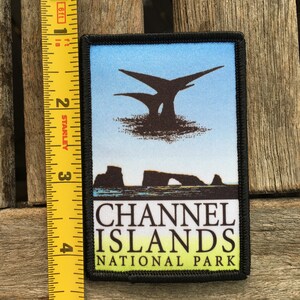 Op de afbeelding: Een geborduurde zwarte en witte patch met een blauwe achtergrond. De patch toont een silhouet van een rotsformatie met een boog en de tekst "CHANNEL ISLANDS NATIONAL PARK".