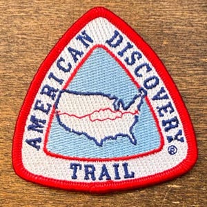 Pode incluir: Remendo bordado triangular com borda vermelha. O remendo apresenta o texto "AMERICAN DISCOVERY TRAIL" em branco, circundando um triângulo azul claro. Dentro do triângulo está um contorno branco dos Estados Unidos com uma linha vermelha.