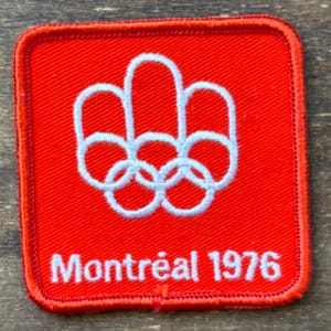 Könnte beinhalten: Ein roter, gestickter Aufnäher mit den olympischen Ringen und einem stilisierten Design darüber. Der Text "Montréal 1976" ist darunter in Weiß gestickt. Der Aufnäher hat einen roten Rand.
