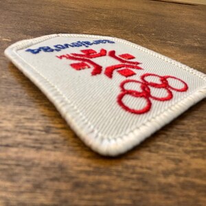 Puede incluir: Un parche rectangular con el texto "Sarajevo 84" en azul, anillos ol&iacute;mpicos rojos y un dise&ntilde;o estilizado. El parche tiene un borde blanco y est&aacute; sobre una superficie de madera.