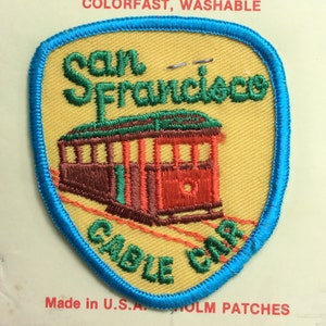Puede incluir: Parche bordado con un tranvía de San Francisco. El parche tiene un fondo amarillo con un borde azul. El texto "San Francisco Cable Car" es verde. El tranvía es rojo y marrón.