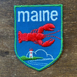 May include: A shield-shaped embroidered patch with a blue background and a bright green border. The word "maine" is stitched in white letters at the top. Below, a red lobster is centered above a white lighthouse on a green grassy hill.