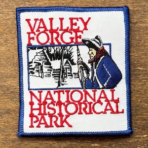 Può includere: Toppa ricamata con sfondo bianco e bordo blu. La toppa presenta il testo "VALLEY FORGE NATIONAL HISTORICAL PARK" in rosso. Un'illustrazione di un soldato e di edifici è al centro.