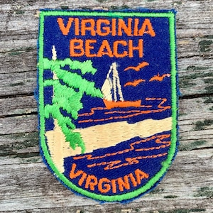 May include: Embroidered patch featuring the words "VIRGINIA BEACH VIRGINIA" in orange on a blue background. The patch depicts a sailboat on the ocean, palm trees, and flying birds. The patch has a green border and is on a wooden surface.