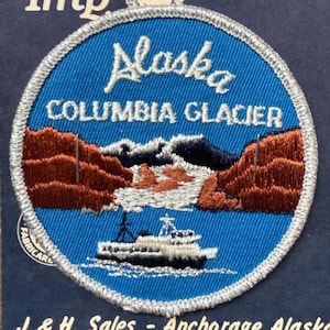 May include: Embroidered patch featuring a blue circle with the word "Alaska" in white script. The patch depicts a landscape with mountains, a glacier, and a boat. The text "COLUMBIA GLACIER" is also present.