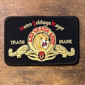 Puede incluir: Un parche rectangular bordado en negro con el logotipo de Metro Goldwyn Mayer. El diseño incluye una cabeza de león central rodeada por un anillo dorado con el texto Ars Gratia Artis, flanqueado por cintas doradas y las palabras Trade Mark.