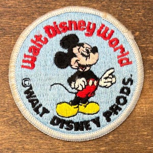 May include: A circular embroidered patch with a light blue background featuring a classic Mickey Mouse character in black, red, and yellow. The text Walt Disney World is stitched in red, with Walt Disney Prods. in black along the bottom edge.