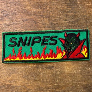 May include: A rectangular embroidered patch with a green background and a black border. The text SNIPES is stitched in black letters. Below the text are red and yellow flames. On the right side is a green devil figure with red horns and a red cape.