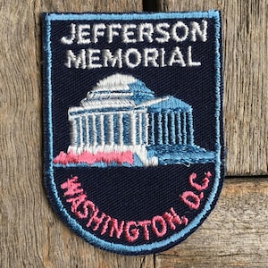 May include: A rectangular embroidered patch with rounded bottom corners featuring a dark blue background. It displays the white text JEFFERSON MEMORIAL above a light blue and white depiction of the monument, with pink text WASHINGTON, D.C. below.