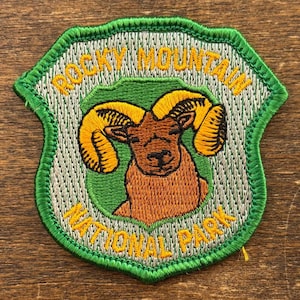 Può includere: Toppa ricamata con il logo del Rocky Mountain National Park. La toppa ha la forma di uno scudo con un bordo verde e uno sfondo grigio chiaro. Il design include un ariete con grandi corna gialle e il nome del parco in giallo.
