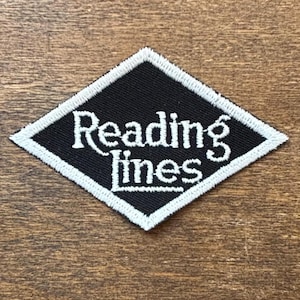May include: A black diamond-shaped patch with white embroidered text that reads "Reading Lines." The patch has a white border and is set against a wooden background. This patch is suitable for jackets, bags, or other fabric items.