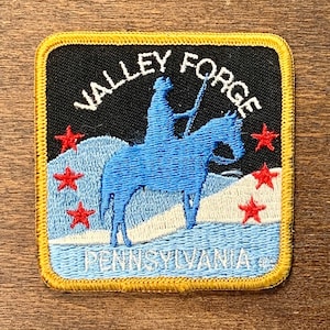 Peut inclure: Un écusson brodé bleu et blanc avec une bordure jaune. Il représente une silhouette d'une personne à cheval avec le texte "Valley Forge Pennsylvania" et cinq étoiles rouges.