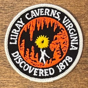 May include: Embroidered patch with a circular design featuring the text "Luray Caverns, Virginia" and "Discovered 1878". The patch has a black background with orange and yellow accents, depicting a cave scene with a person and a flower.