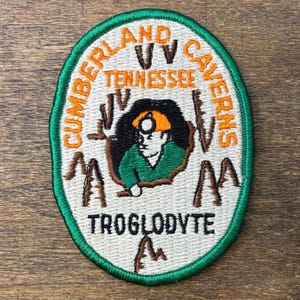 May include: Embroidered patch with a green border and an oval shape. The patch features the text "Cumberland Caverns Tennessee" in orange, surrounding an image of a miner. The word "Troglodyte" is at the bottom.
