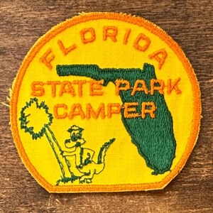 May include: A round, yellow patch with orange text that reads "FLORIDA STATE PARK CAMPER." The patch features a green outline of Florida and a cartoon alligator with a palm tree. The patch has an orange border.