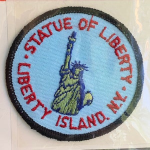 May include: Embroidered patch featuring the Statue of Liberty in green and blue on a light blue background. The text "STATUE OF LIBERTY, N.Y. LIBERTY ISLAND" is embroidered in red around the image, with a black border.