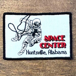 May include: Embroidered patch with a black and white astronaut design, complete with a tether, set against a pale blue backdrop. The words "SPACE CENTER" in red and "Huntsville, Alabama" are also displayed.