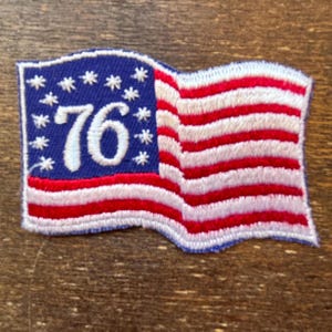 Peut inclure: Écusson brodé représentant un drapeau américain flottant. Le motif comprend un champ bleu avec des étoiles blanches, des rayures rouges et blanches, et le chiffre "76" en blanc. L'écusson est bordé d'une couture blanche.