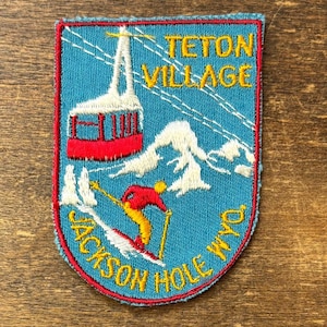May include: A rectangular embroidered patch with a light blue background and a red border. It features a red aerial tram, white snow-capped mountains, and a skier in a red jacket and yellow pants. Text reads TETON VILLAGE and JACKSON HOLE WYO.