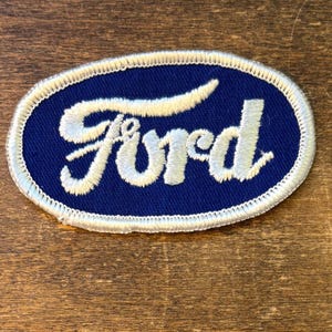 May include: An oval-shaped patch featuring the word "Ford" embroidered in white script on a navy blue background. The patch is outlined with a white stitched border. The patch is likely for a jacket or other apparel.