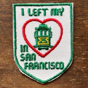 Puede incluir: Parche bordado con fondo blanco, borde verde y el texto "I LEFT MY IN SAN FRANCISCO". Un corazón rojo rodea un tranvía verde. El parche tiene forma de escudo.