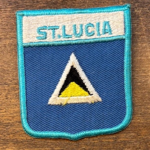 May include: Embroidered patch featuring the text "ST. LUCIA" above a blue shield with a white, black, and yellow triangle design. The patch has a blue border and is set against a wooden surface.