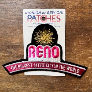 Könnte beinhalten: Gestickter Aufnäher mit dem Wort "RENO" in Rosa und dem Satz "THE BIGGEST LITTLE CITY IN THE WORLD" in Weiß auf schwarzem Hintergrund. Über "RENO" befindet sich ein goldfarbenes Burst-Design.